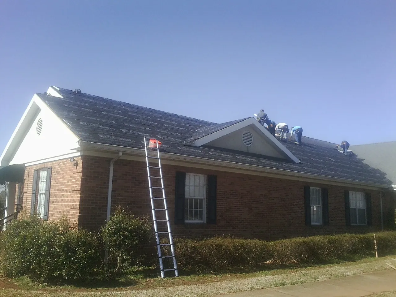 Crew tearing shingles off a brick ranch roof with a ladder set — Emergency Roof Repair work by our crew in Mackinaw