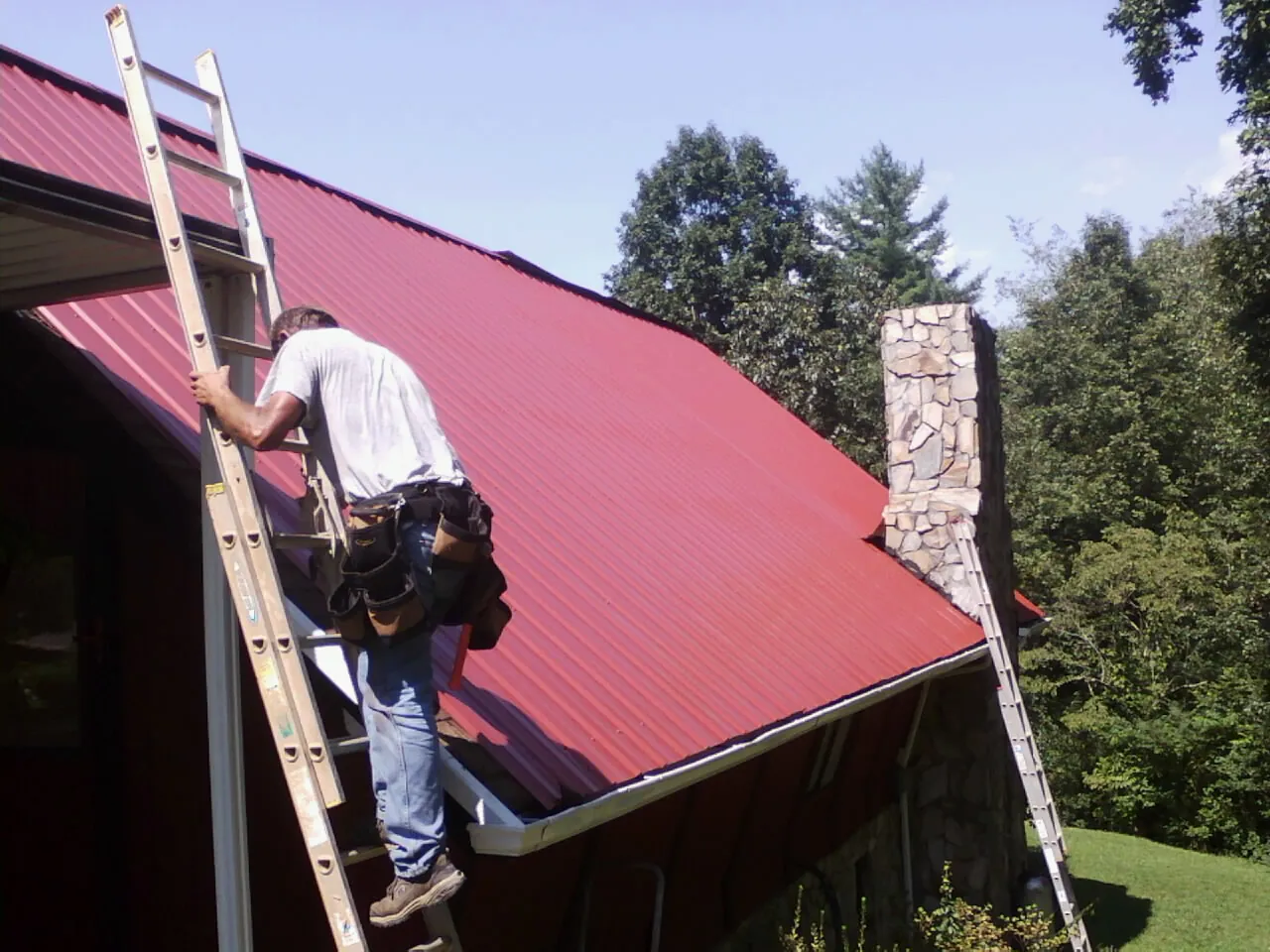 Roofer on a ladder beside a red metal roof and stone chimney — Soffit & Fascia Repair by Roofing Brothers in Mackinaw